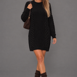 Black cable-knit long-sleeve jumper dress on a model against a grey studio background.