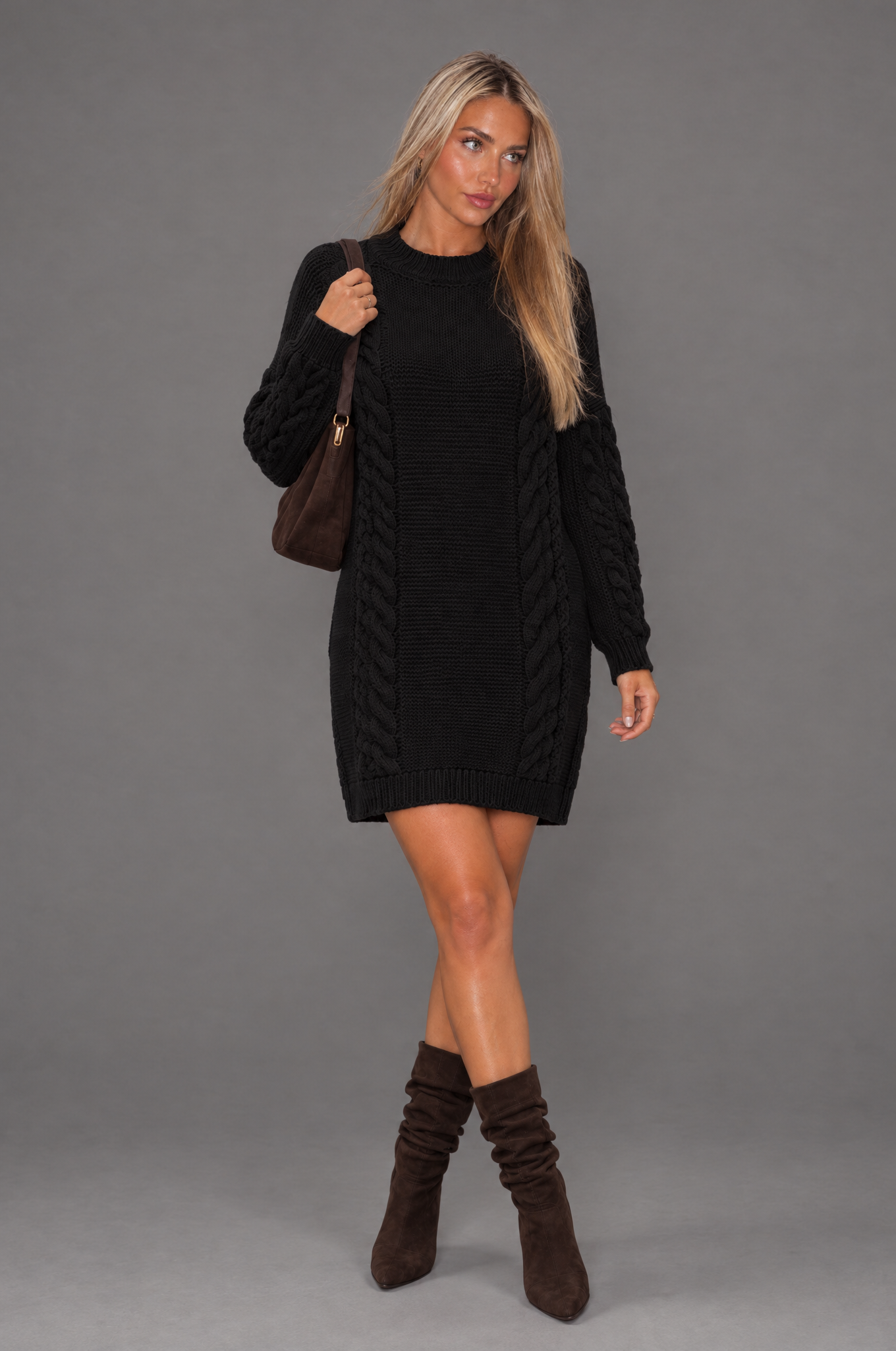 Black cable-knit long-sleeve jumper dress on a model against a grey studio background.