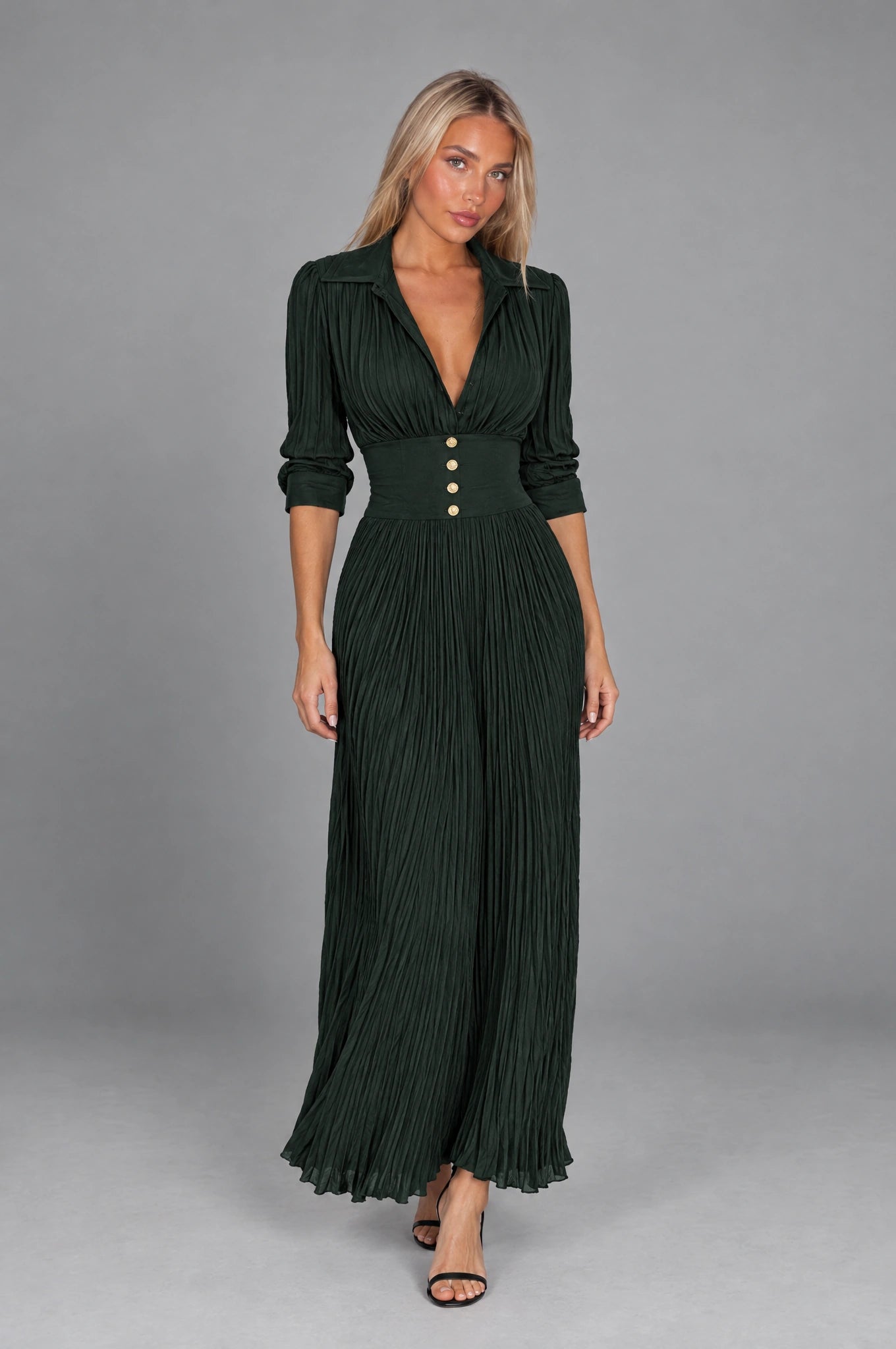 Dua York model wearing the dark-green Elvira pleated maxi dress with four gold buttons against a grey studio background